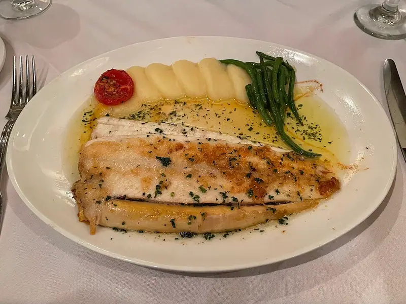 Pan-fried sole fillets in a herb-flecked butter sauce with piped mashed potato, green beans and a roasted tomato half on an oval plate
