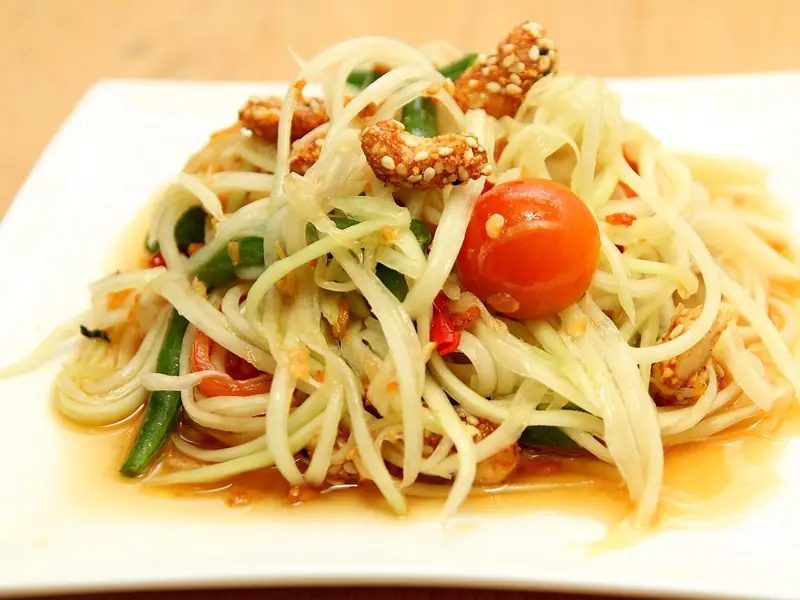 Shredded green papaya salad with long beans, a cherry tomato, red chilli and sesame-coated cashews in a light dressing on a white plate.
