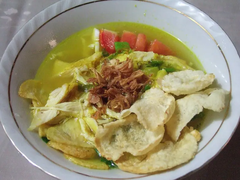 Bowl of yellow turmeric chicken soup with shredded chicken, tomato wedges, spring onion, fried shallots and puffy crackers on top.