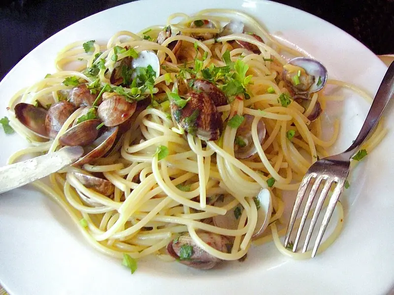 Spagetti apró, héjas vongole kagylóval és aprított petrezselyemmel fehér tányéron, a tányér szélén villával és késsel.