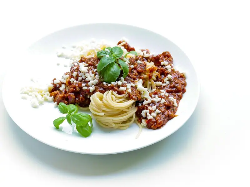 Ein Spaghettinest mit Fleischsauce, geriebenem weißem Käse und frischen Basilikumzweigen auf einem weißen Teller vor weißem Hintergrund.