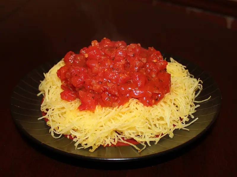 Dampfende Spaghettikürbis-Fäden auf einem dunklen Teller, darauf ein Berg stückiger Tomatensauce mit Kräutern und Chiliflocken.