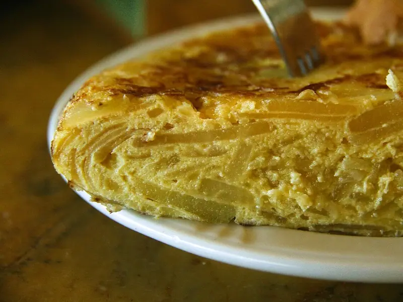 Beyaz bir tabakta altın rengi, kalın bir dilim patatesli ve soğanlı omlet; çatal kızarmış üst kısmını kesiyor.