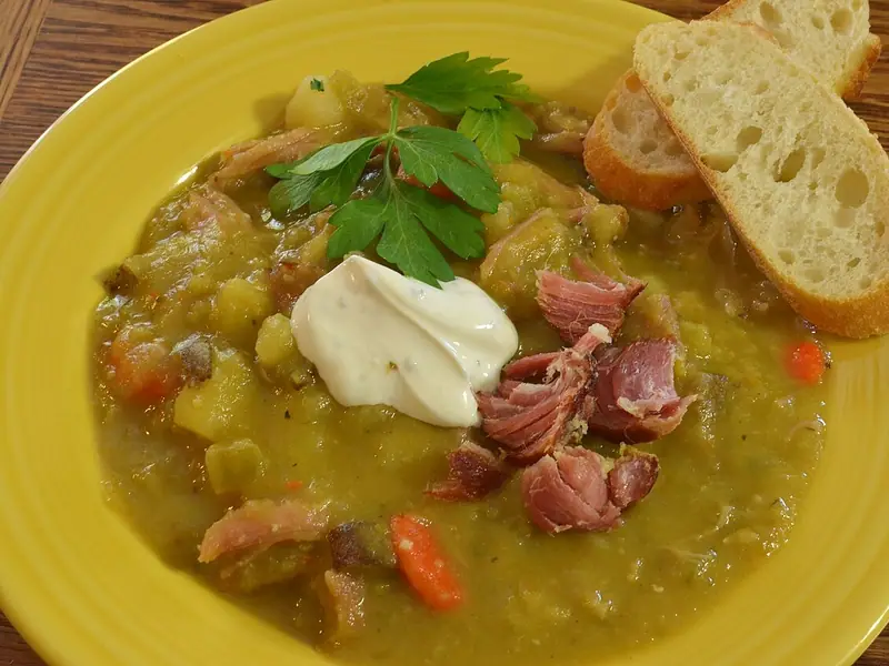 Chunky split pea soup on a yellow plate topped with shredded ham, a dollop of sour cream and parsley, with two baguette slices