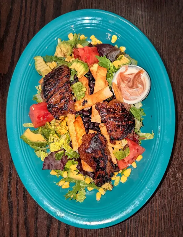 Grilled steak pieces over greens with black beans, corn, avocado, tomato wedges and tortilla strips, with a creamy dip, on a teal plate
