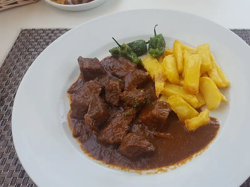 Rindfleischstücke in dunkelbrauner Sauce mit dick geschnittenen Pommes frites und angerösteten grünen Paprikaschoten auf einem weißen Teller, daneben Messer und Gabel.