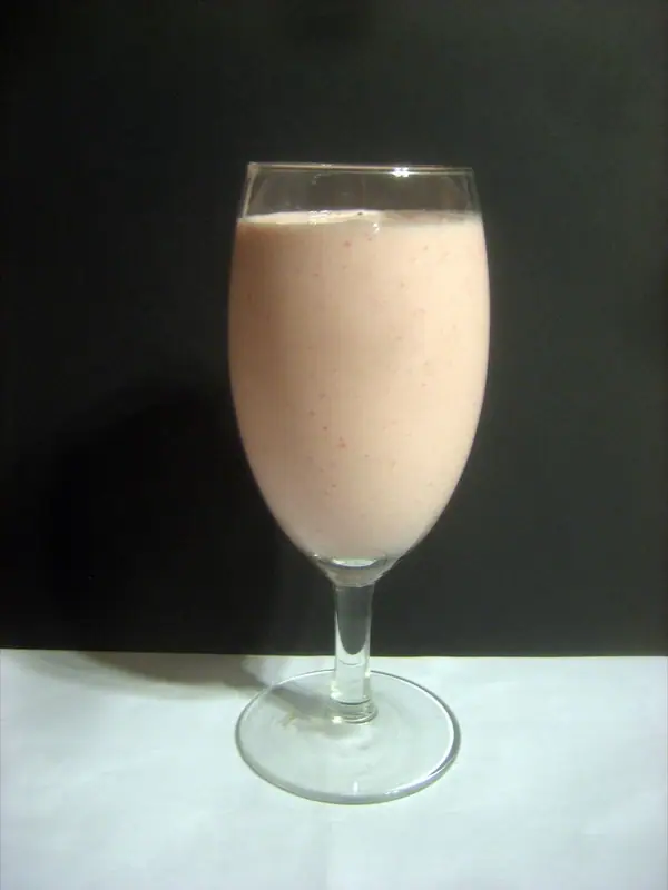 Ένα απαλό ροζ smoothie με μικρές κόκκινες κουκκίδες σε ποτήρι κρασιού με πόδι, πάνω σε λευκή επιφάνεια με μαύρο φόντο.