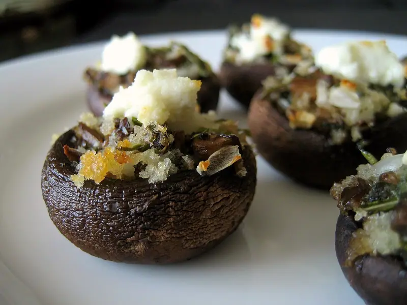 Baked brown mushroom caps stuffed with herby breadcrumb filling and topped with crumbled white cheese on a white plate.