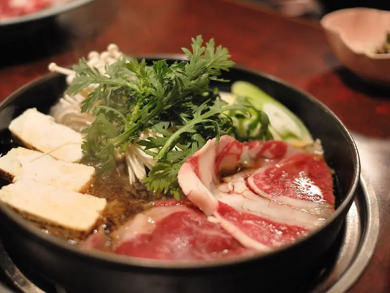 Oală neagră de sukiyaki pe un reșou de masă, cu felii crude de vită marmorată, cuburi de tofu, ciuperci enoki, praz și crizantemă comestibilă proaspătă.