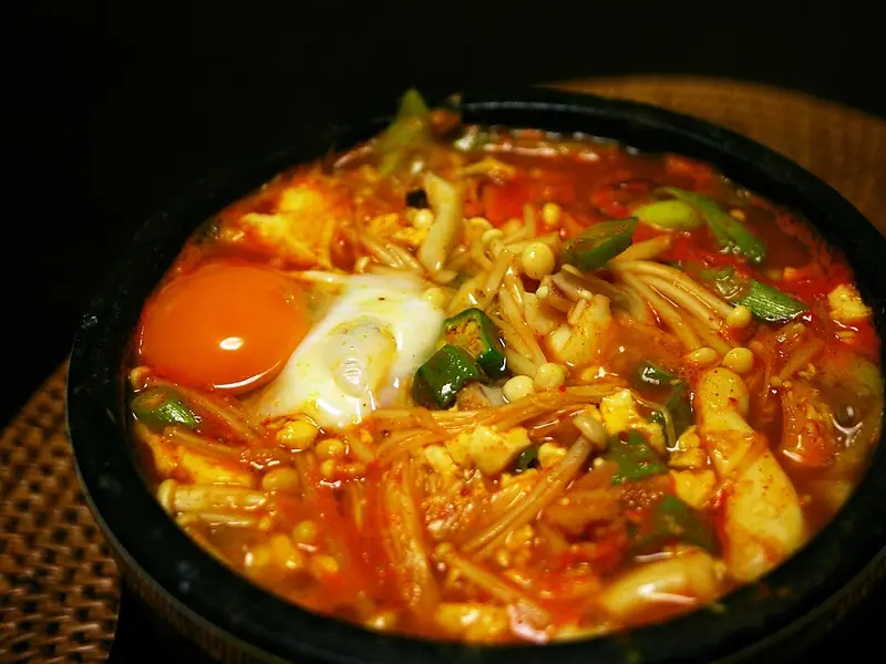 Red spicy soft tofu stew in a black stone pot with a raw-looking egg yolk, enoki mushrooms, green chilli and scallions on a wicker mat.