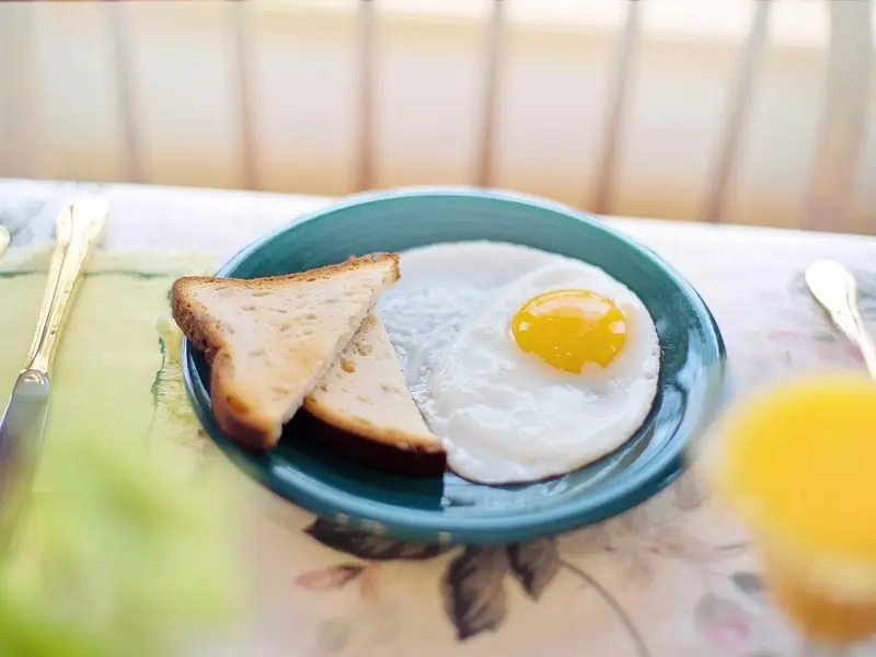 Jedno volské oko vedle dvou trojúhelníků toastu na modrozeleném talíři, s příborem a pomerančovým džusem na květovaném ubrusu.