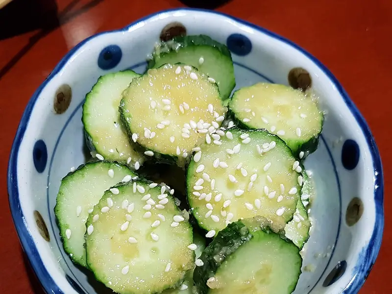 Thick cucumber rounds glossed with a pale ginger dressing and sprinkled with sesame seeds in a blue polka-dot bowl on a red table.