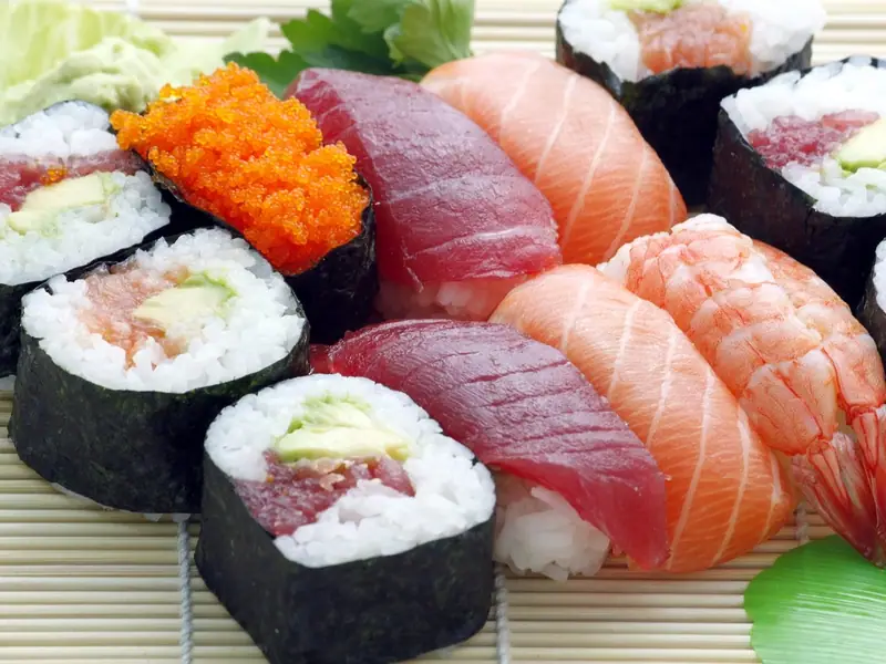 Salmon and tuna nigiri, a shrimp nigiri, orange roe gunkan and avocado maki rolls arranged on a bamboo mat with green garnish.