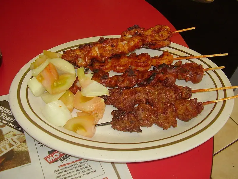 Five grilled meat skewers with reddish glaze, some chicken and some beef, beside chunks of raw onion and tomato on an oval plate