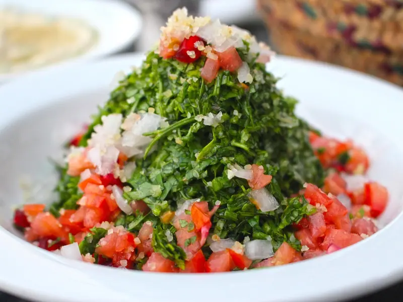 A white bowl piled high with finely chopped parsley tabbouleh, ringed with diced tomato and topped with chopped white onion and bulgur.