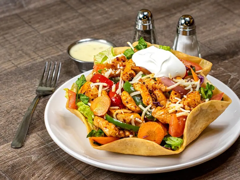 Krydrede kyllingestrimler, peberfrugt, gulerødder, tomater og ost i en sprød tortillaskål med creme fraiche på toppen og dressing ved siden af.