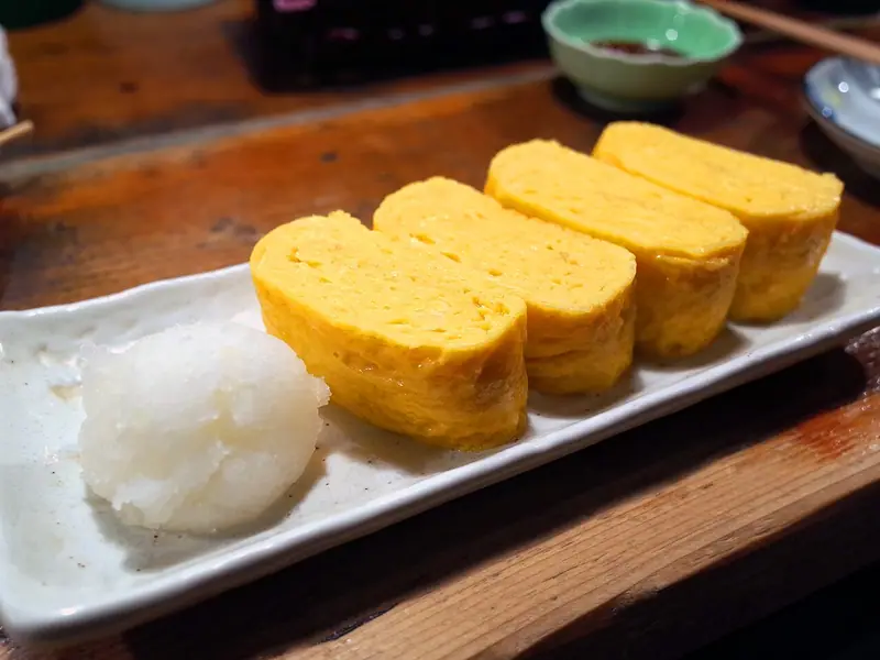 Four thick slices of golden rolled Japanese omelette on a white rectangular plate beside a mound of grated daikon radish.