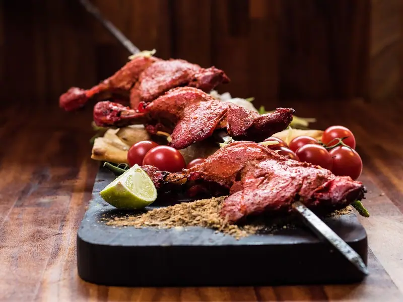 Bright red tandoori chicken legs on skewers on a dark board with cherry tomatoes, a lime wedge, ground spices and flatbread