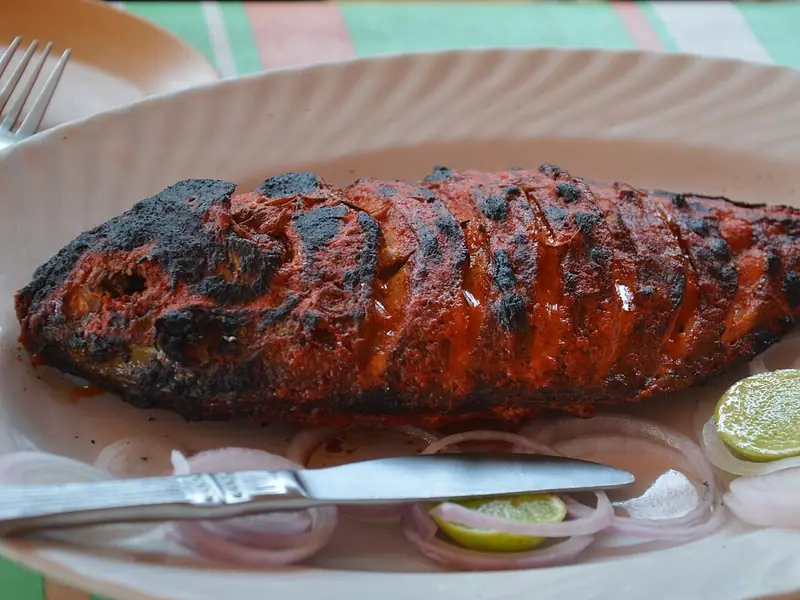 Celá naříznutá ryba s opečenou červenou tandoori krustou na bílém talíři, vedle syrové cibulové kroužky a půlky limetky u nože a vidličky.