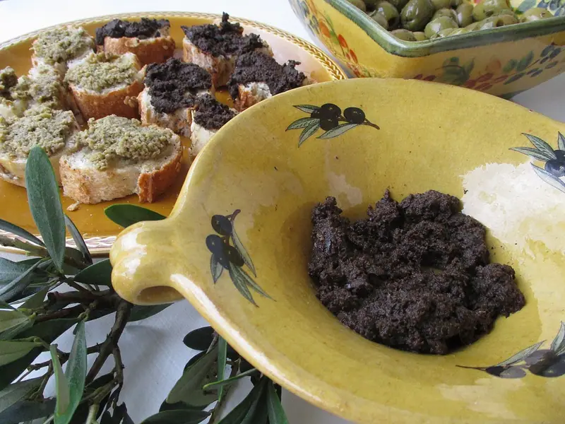 Dark black olive tapenade in a yellow olive-painted dish, with black and green tapenade on bread slices and a bowl of green olives behind