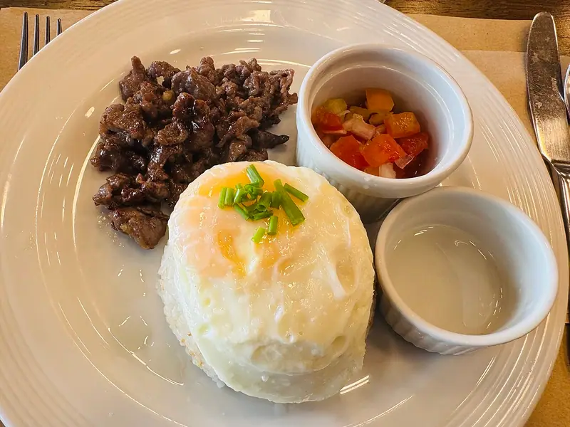 Seared beef tapa beside a mound of rice topped with a fried egg and chives, with small cups of diced tomato salsa and vinegar.