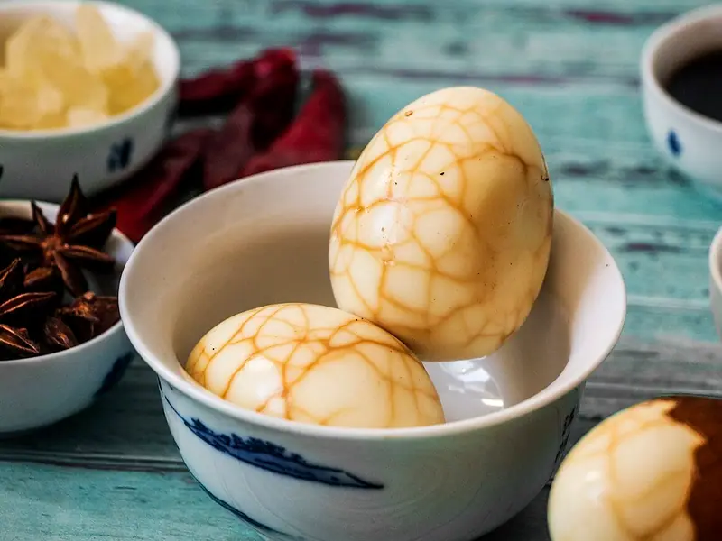 Two peeled marbled tea eggs in a white blue-patterned cup, with star anise, rock sugar, dried chilies and soy sauce on a teal table.