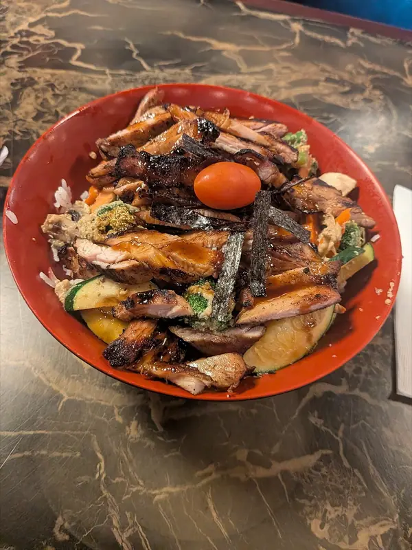 Poulet teriyaki grillé et tranché sur du riz blanc, avec courgette, brocoli et carottes dans un bol rouge, garni de nori et d'une tomate cerise.