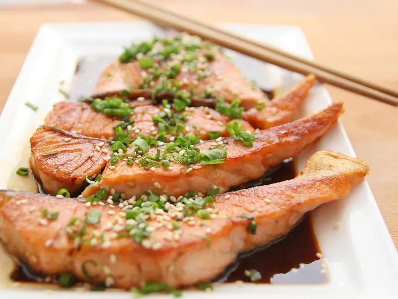 Glazed seared salmon pieces topped with sliced spring onions and sesame seeds in dark sauce on a white plate, chopsticks resting on top