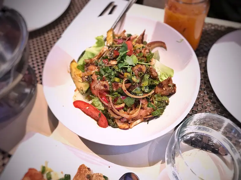Thailandsk salat med grillet kjøtt, rødløk, tomat, koriander og salat i en hvit bolle, med en oransje drikk ved siden av på bordet.