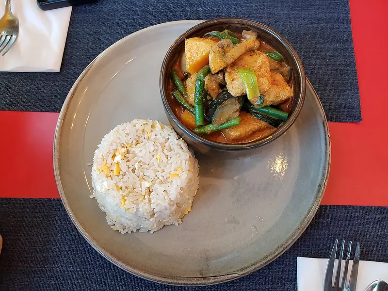 Red coconut curry with fried tofu, squash, green beans and mushrooms in a dark bowl beside a mound of egg-fried rice