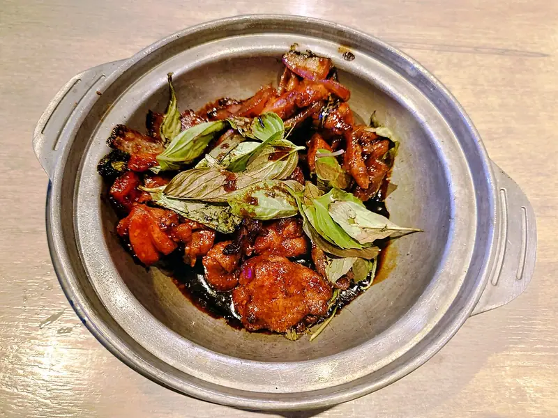 Glossy dark-braised chicken pieces topped with fresh basil leaves, served in a round metal clay-pot-style dish on a wooden table.
