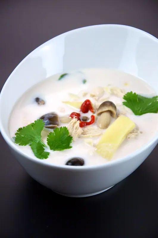 Cremet hvid kokossuppe i en hvid skål med revet kylling, halmsvampe, bambusskud, rød chili og friske korianderblade.