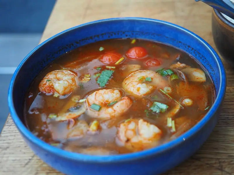 Blue bowl of reddish-orange broth with shrimp, cherry tomatoes, mushrooms, lemongrass and chopped coriander on a wooden table.