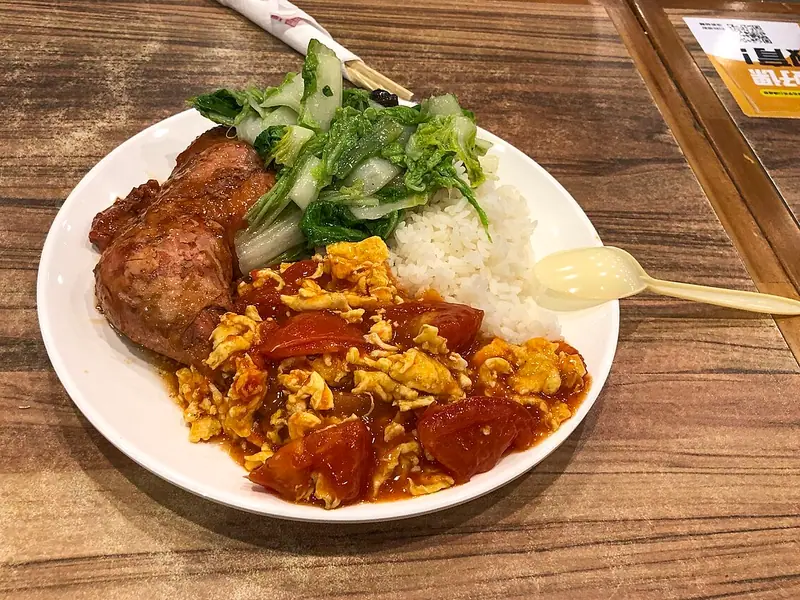 木のテーブルに置かれた、トマトと卵の炒め物、白ごはん、白菜炒め、照りのある鶏もも肉の煮込みを盛り付けたプレート。