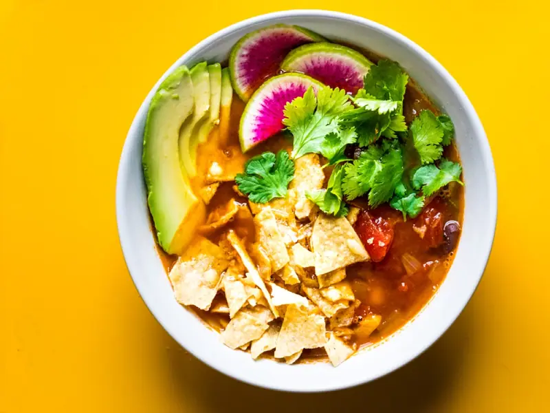 Hvid skål med orangerød tortillasuppe toppet med tortillachips, avocadoskiver, vandmelonradise og koriander på et gult bord.
