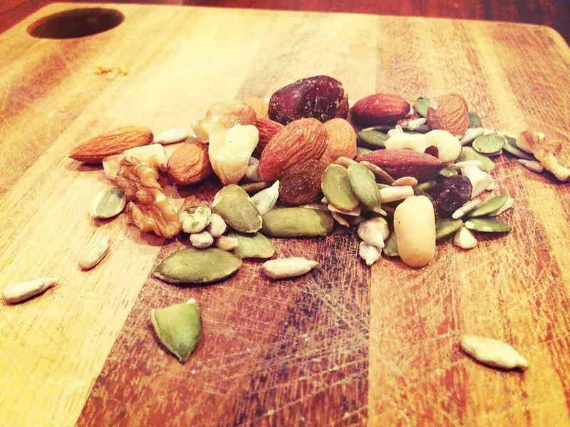 A loose pile of almonds, walnuts, pumpkin seeds, sunflower seeds, peanuts and raisins scattered on a wooden cutting board.