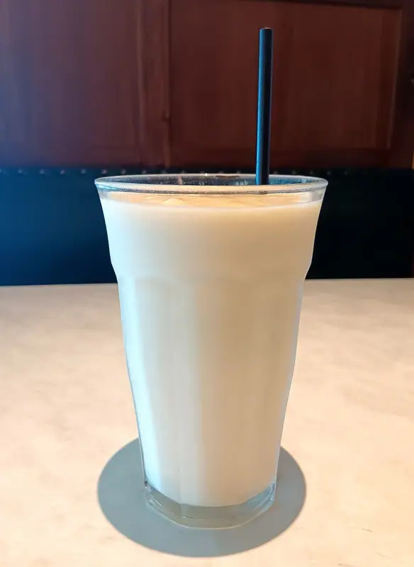 Vysoká sklenice světlého, krémově bílého smoothie s černým brčkem na světlém mramorovém stole před boxem z tmavého dřeva.