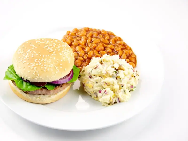 Burger într-o chiflă cu susan, cu salată verde și ceapă roșie, servit pe o farfurie albă alături de fasole la cuptor și salată cremoasă de cartofi.