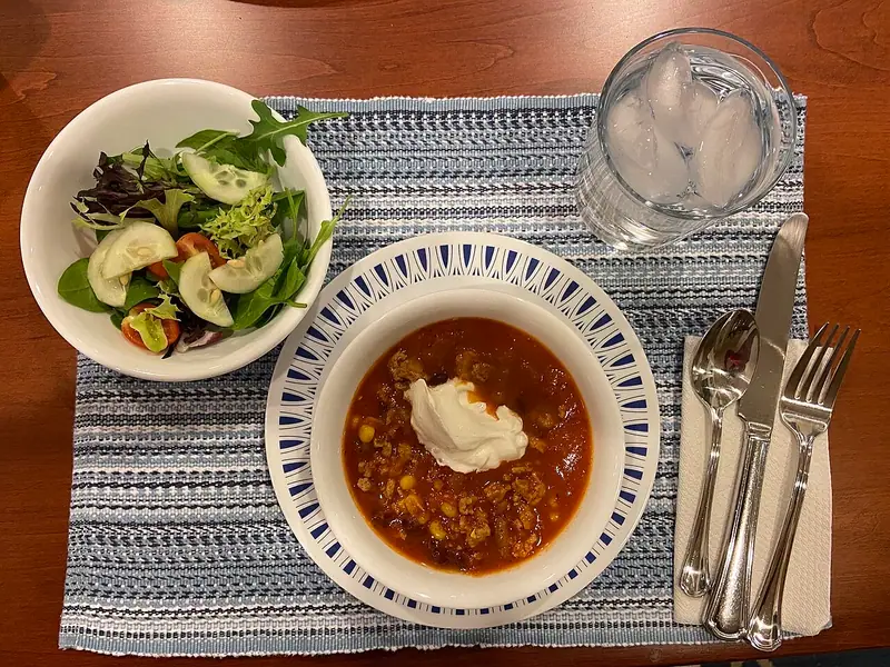 Tigela de chili de peru com milho e uma colherada de creme azedo, ao lado de uma salada verde com pepino, um copo de água gelada e talheres.