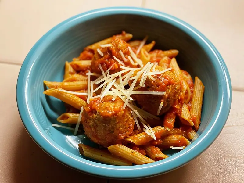 Meatballs in tomato sauce over penne pasta, topped with shredded Parmesan, in a turquoise bowl on a beige tiled surface.