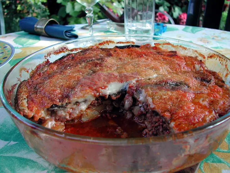Round glass dish of baked moussaka with a browned cheese topping, a portion scooped out to show meat and potato layers, on a floral table