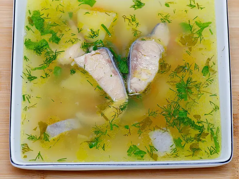 Clear golden fish soup in a square white bowl with skin-on fish steaks, potato chunks and lots of chopped dill and parsley on top.