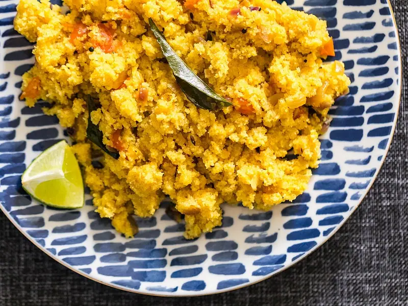 Yellow, crumbly semolina upma with tomato bits, onion and curry leaves on a blue and white patterned plate with a lime wedge.
