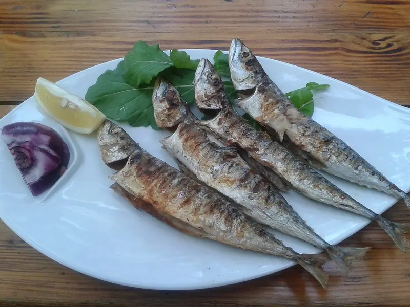 Fire hele grillede makreller med sprødt, grillsværtet skind på et hvidt ovalt fad med rucola, en citronbåd og et kvart rødløg på et træbord.