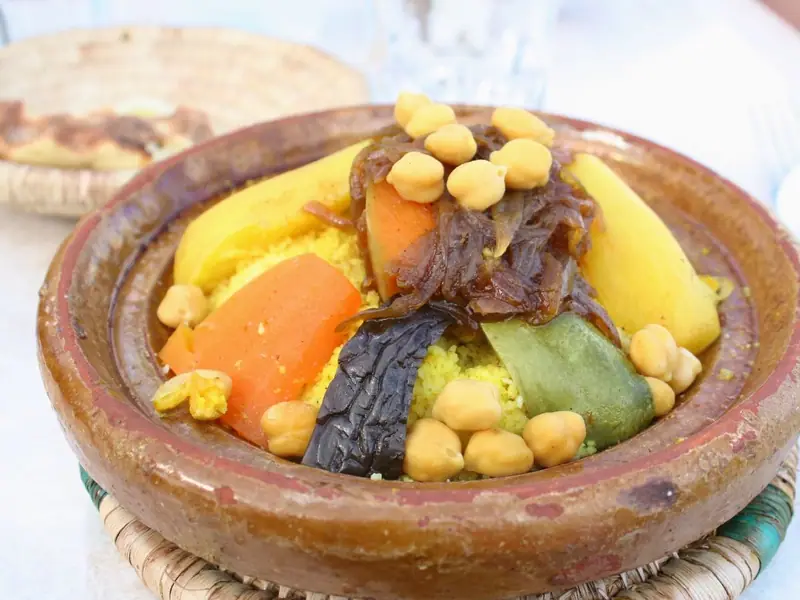 Couscous i et glaseret lerfad med gulerødder, græskar, aubergine, grøn peberfrugt, kikærter og karamelliserede løg, med brød i baggrunden.