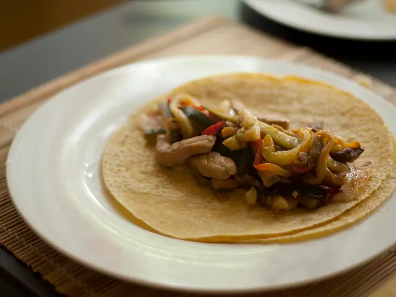 A soft tortilla on a white plate topped with strips of meat, yellow and red peppers, zucchini and onion, set on a bamboo placemat.