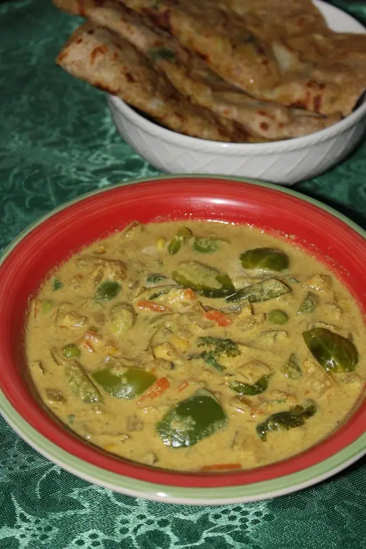 Creamy yellow vegetable curry with green pepper, Brussels sprouts, carrot and peas in a red bowl, with flatbreads in a white bowl behind