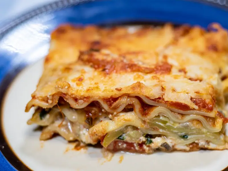 Gruby kawałek warstwowej lasagne z sosem pomidorowym, ricottą, cukinią i szpinakiem pod złocistą zapieczoną warstwą sera, na talerzu z niebieskim brzegiem.