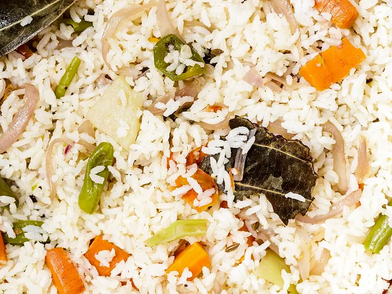 Close de pulao de arroz branco com folhas de louro, pimentas verdes, cebola fatiada, cenoura em cubos, vagem e pedaços de repolho