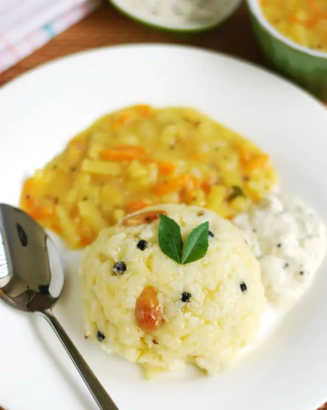 En kuppel af cremet ven pongal med hele sorte peberkorn, en cashewnød og karryblade på en hvid tallerken med grøntsagssambar og kokoschutney